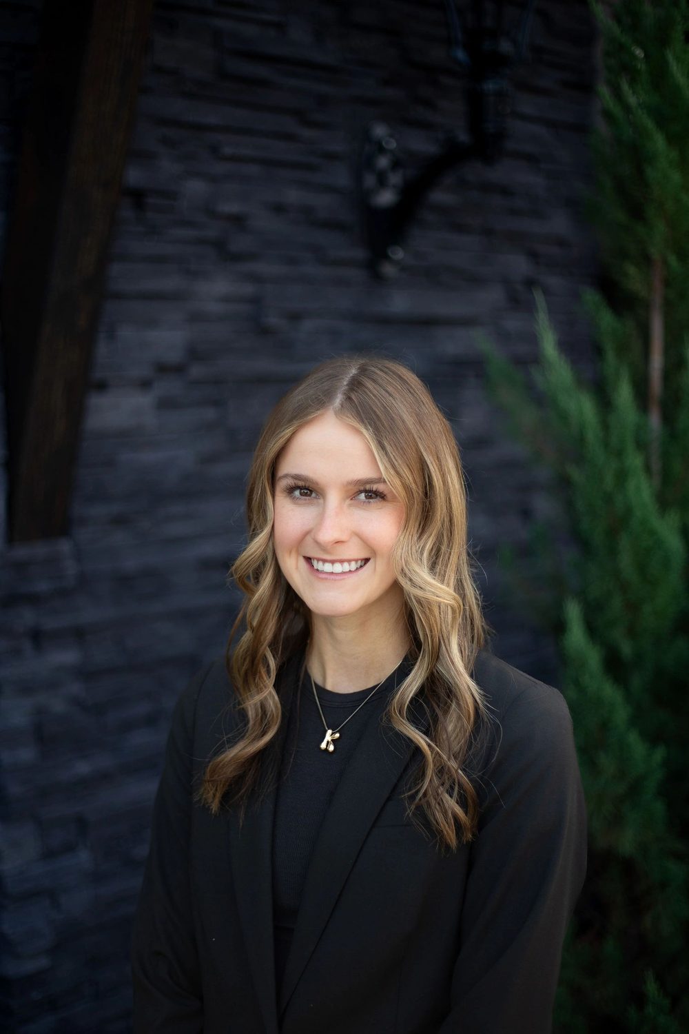 Kinley Rening smiling in a black blazer in front of a dark stone wall and evergreen trees